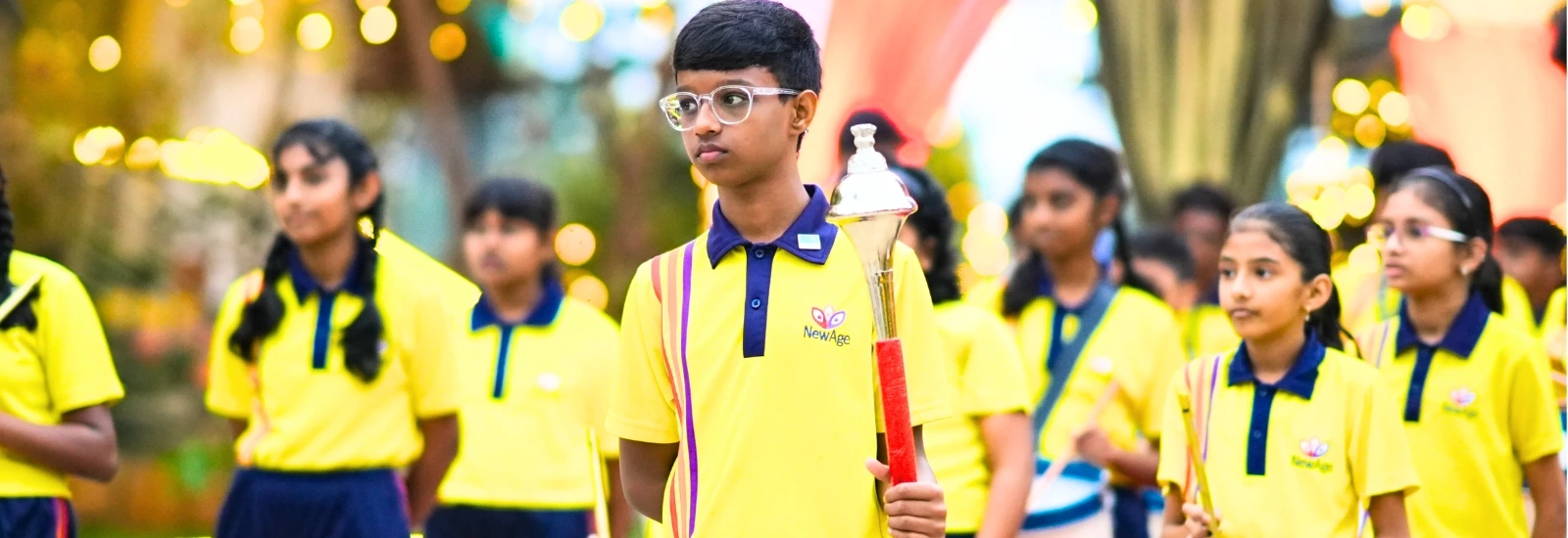 Students of NewAge World School participating in a ceremonial march, dressed in school uniforms, with a student leading the procession holding a torch.